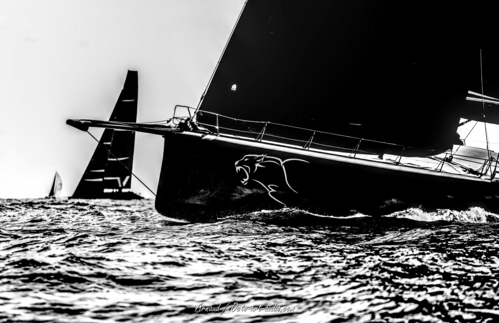 Gros plan artistique en noir et blanc de l'étrave du Maxi Yacht Leopard 3 en pleine mer, logo de léopard visible sur la coque sombre, avec un autre voilier en arrière-plan lors des Voiles de Saint-Tropez 2025.