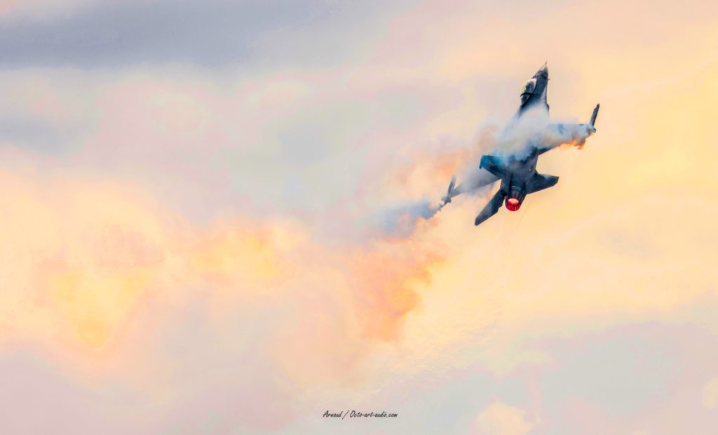 Photographie d'art d'un avion de chasse F-16 Fighting Falcon en manœuvre ascendante avec postcombustion allumée, traversant des nuages de condensation aux teintes dorées.