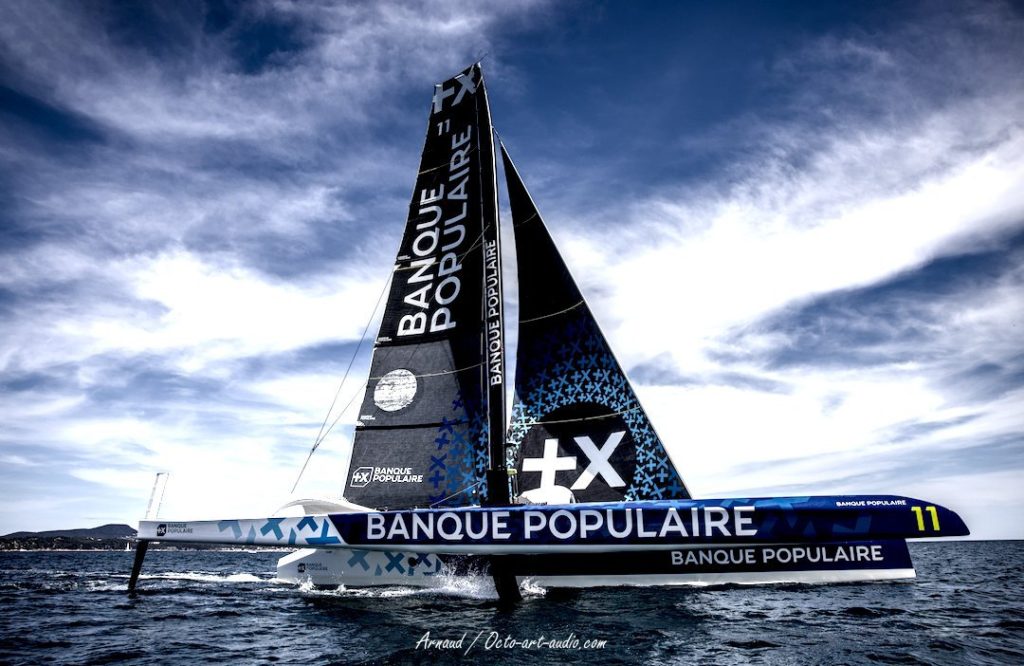 Le trimaran Ultim Banque Populaire XI volant sur ses foils au large, ciel contrasté et design bleu et blanc emblématique.