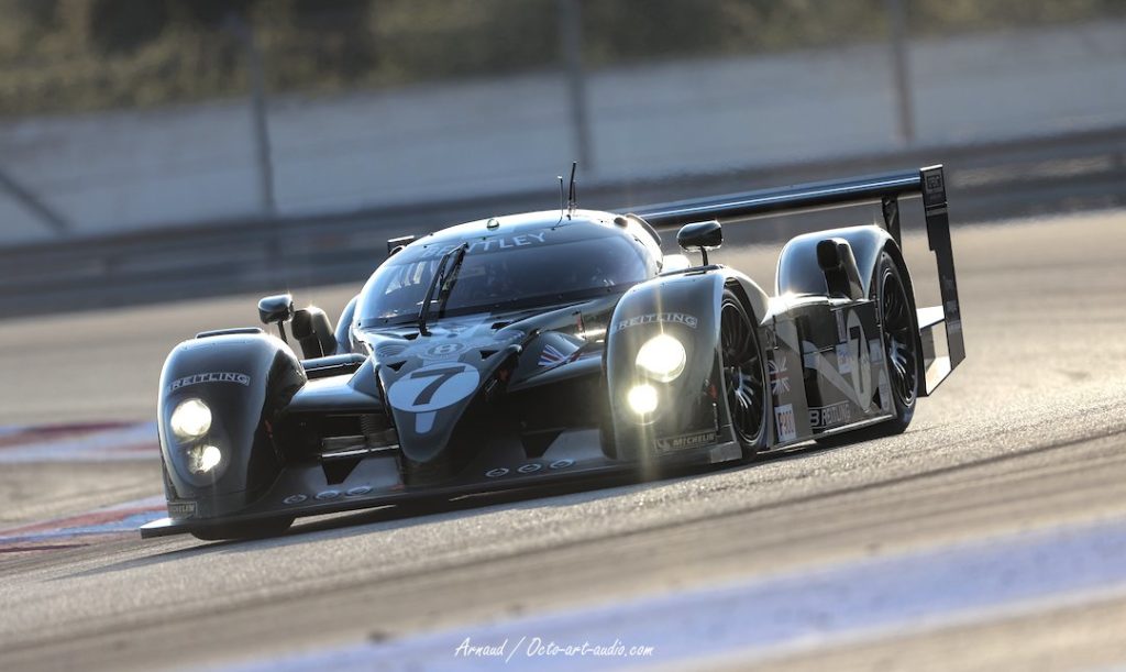 Photographie d'art d'un prototype Bentley Speed 8 (numéro 7) sur circuit du Paul Ricard, avec les phares allumés et des reflets de lumière sur sa carrosserie vert "British Racing Green".