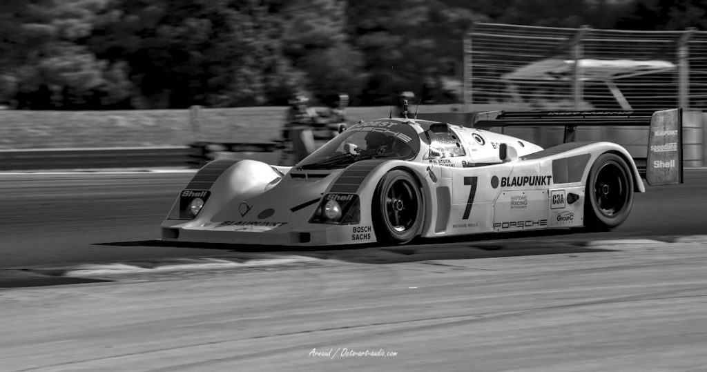 Photographie artistique en noir et blanc d'une Porsche 962 de course (livrée Blaupunkt) en pleine action sur circuit lors des dix mille tours Peter Auto.