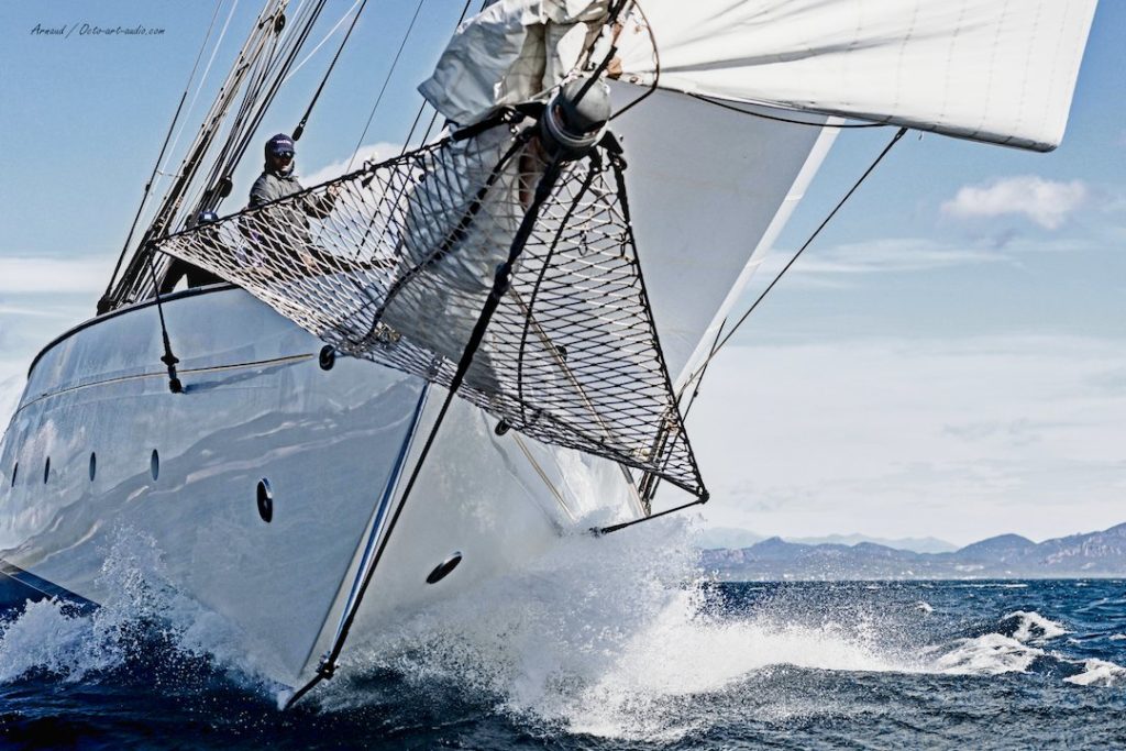 Vue impressionnante de l'étrave du voilier Naema fendant les vagues lors des Voiles de Saint-Tropez, éclats d'écume et voiles blanches.