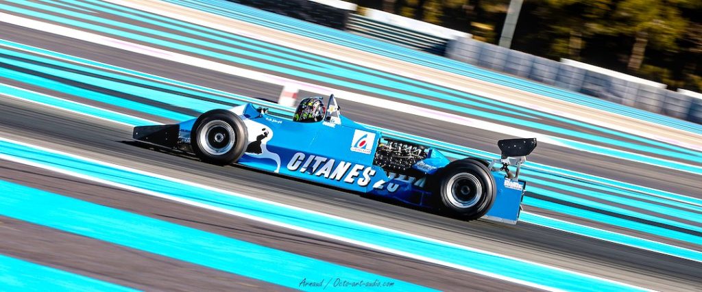 Photographie en filé dynamique de la Formule 1 Ligier JS11 bleue Gitanes sur le circuit Paul Ricard, angle incliné et couleurs vives.