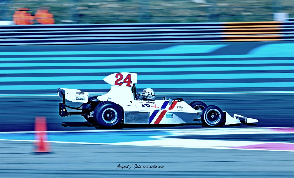 Photographie en filé parfait de la F1 historique Hesketh 308 n°24 blanche de James Hunt gîtant sur le circuit Paul Ricard, couleurs vibrantes.