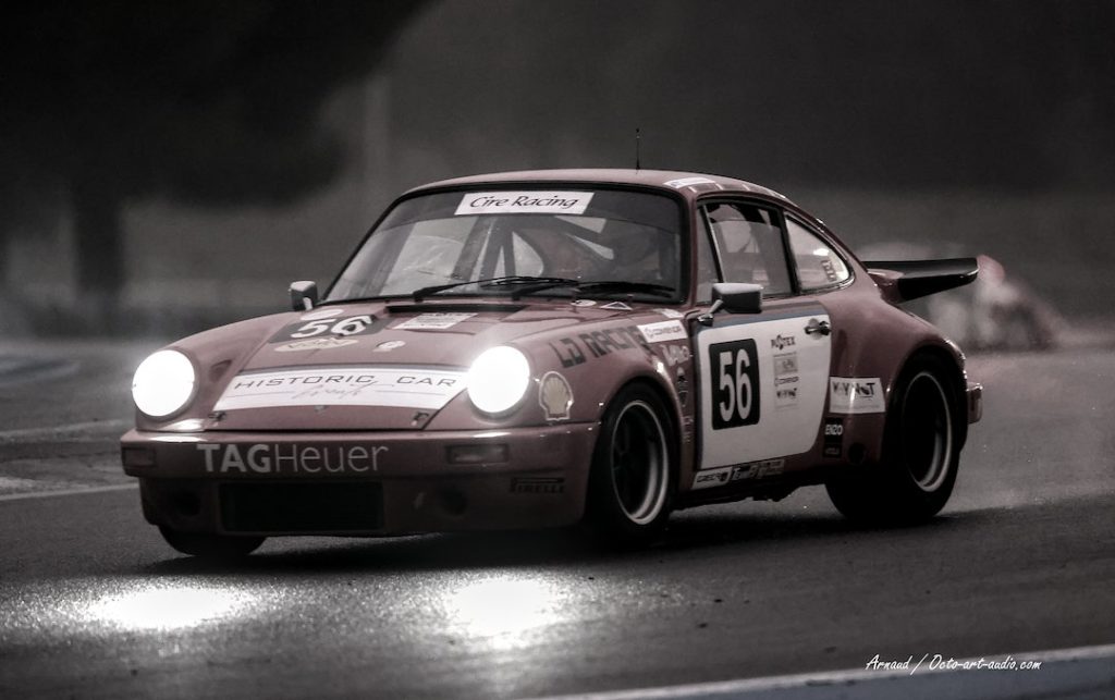 Photographie d'art vintage d'une Porsche 911 de course historique (numéro 56) de nuit sur piste mouillée, phares allumés et sponsors TAG Heuer visibles.