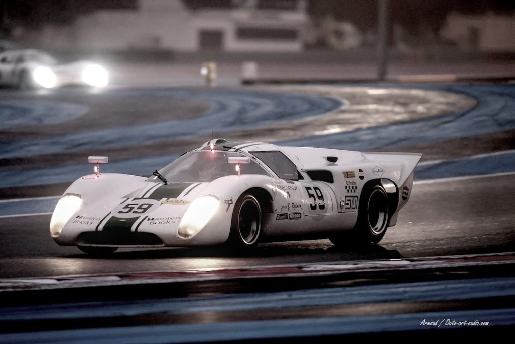 Prototype Lola T70 MkIII numéro 59 en course de nuit sur piste mouillée au circuit Paul Ricard, phares allumés et reflets sur le bitume.