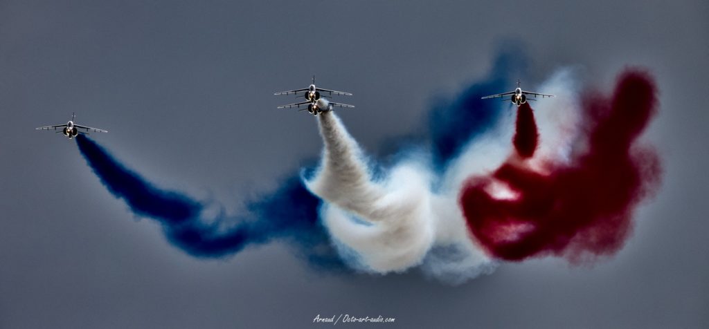 Gros plan artistique de quatre Alpha Jets de la Patrouille de France volant en formation, chacun laissant un panache de fumée de couleur : bleu, blanc, rouge, dessinant un drapeau français dans le ciel. La perspective est d'en dessous, cadrée sur l'avant des appareils et les traînées colorées.