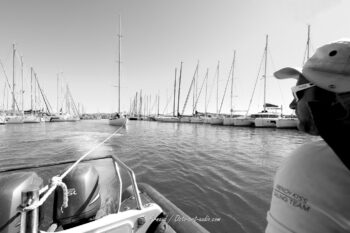 Mise en place matinale. Un membre de la French Kiss Sailing Team supervise le remorquage du 12mJI South Australia à travers le bassin du port de Hyères lors de l'Europa Cup 2026. Tirage d'art disponible en édition limitée. Photographie en noir et blanc du remorquage du voilier 12mJI South Australia par l'équipe d'assistance de French Kiss Sailing Team quittant le port de Hyères lors de l'Europa Cup 2026.