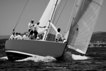 L'effort et l'élégance. Le mythique 12mJI France 1 s'appuie sur son étrave sous les manœuvres coordonnées de ses équipiers lors de l'Europa Cup 2026. Tirage d'art disponible en édition limitée. Photographie d'art en noir et blanc de trois-quarts avant du voilier classique 12mJI France 1 fendant les vagues avec son équipage en action, lors de l'Europa Cup 2026 à Hyères.
