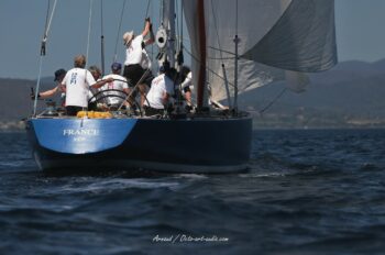 Manœuvre de portant à bord de France 1. L'équipage du mythique 12mJI règle le spinnaker face aux reliefs du Var lors de l'Europa Cup 2026. Tirage d'art disponible en édition limitée. Photographie de trois-quarts arrière du voilier de tradition 12mJI France 1 à coque bleue, montrant l'équipage en action lors de la manœuvre du spinnaker pendant l'Europa Cup 2026 à Hyères.