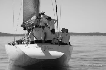 Concentration à la poupe. L'équipage de French Kiss (F-12) fait corps à l'arrière du célèbre 12mJI français lors d'une phase calme de l'Europa Cup 2026. Tirage d'art disponible en édition limitée. Photographie d'art en noir et blanc du tableau arrière du voilier classique 12mJI French Kiss F-12 avec son équipage de dos portant des t-shirts F-7 lors de l'Europa Cup 2026 à Hyères.