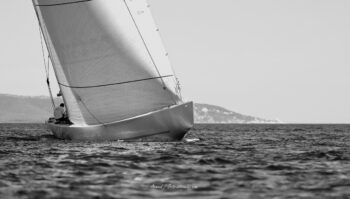 Perspective classique en haute mer. La silhouette graphique du 12mJI French Kiss (F-12) s'efface vers l'horizon lors des régates de l'Europa Cup 2026. Tirage d'art disponible en édition limitée. Photographie d'art en noir et blanc du voilier de tradition 12mJI French Kiss F-12 naviguant de trois-quarts arrière, vue sur le pont et la grand-voile déployée lors de l'Europa Cup 2026 à Hyères.