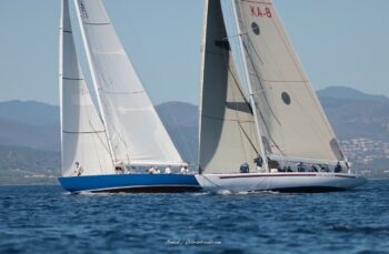 Duel au sommet en rade de Hyères. Les mythiques 12mJI France 1 et South Australia (KA-8) croisent au plus près lors des régates tactiques de l'Europa Cup 2026. Tirage d'art disponible en édition limitée. Photographie de profil montrant le croisement serré entre les voiliers de tradition 12mJI France 1 (coque bleue) et South Australia KA-8 (coque blanche) lors de l'Europa Cup 2026 à Hyères.