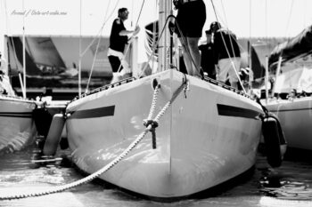 Graphisme et puissance à quai. Focus sur la ligne de proue du 12mJI South Australia (KA-8) dans le bassin du port de Hyères pendant l'Europa Cup 2026. Tirage d'art disponible en édition limitée. Photographie d'art en noir et blanc de l'étrave et du pont du voilier classique 12mJI South Australia KA-8 amarré à quai avec son équipage lors de l'Europa Cup 2026 à Hyères.