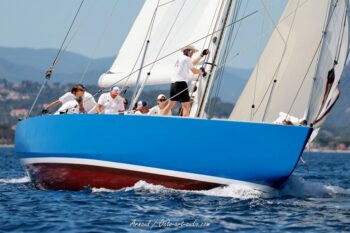 Puissance au ras des vagues. L'étrave du mythique 12mJI France 1 fend la Méditerranée sous les manœuvres de son équipage lors de l'Europa Cup 2026. Tirage d'art disponible en édition limitée. Photographie de l'étrave du voilier classique 12mJI France 1 à coque bleue et rouge fendant l'eau avec son équipage en action, lors de l'Europa Cup 2026 à Hyères.