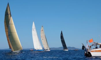 Tension sur la ligne de départ. La flotte internationale des 12mJI s'élance sous le contrôle du bateau comité traditionnel lors de l'Europa Cup 2026 à Hyères. Tirage d'art disponible en édition limitée. Photographie de l'alignement des voiliers classiques 12mJI lors d'un départ de course, franchissant la ligne à côté du bateau comité traditionnel lors de l'Europa Cup 2026 à Hyères.