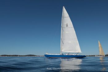 Sérénité et lignes pures. Le légendaire 12mJI France 1 glisse sur les ondulations de la rade de Hyères lors de l'Europa Cup 2026. Tirage d'art disponible en édition limitée. Photographie de profil du voilier classique 12mJI France 1 à coque bleue naviguant sur une mer calme avec de légères ondulations lors de l'Europa Cup 2026 à Hyères.