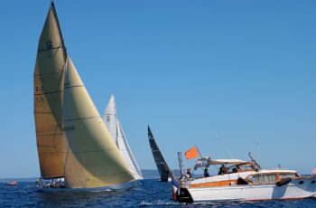 Sous les ordres du comité. Le 12mJI France (F-7) coupe la ligne de départ sous le contrôle du bateau comité traditionnel lors de l'Europa Cup 2026 à Hyères. Tirage d'art disponible en édition limitée. Photographie du départ de course des voiliers classiques 12mJI, montrant le voilier France F-7 sous voiles ambrées franchissant la ligne devant le bateau comité en bois verni lors de l'Europa Cup 2026 à Hyères.
