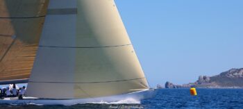 Cap sur la marque de parcours. Le 12mJI French Kiss (F-12) et sa voilure ambrée virent devant le rocher du Crocodile à Porquerolles lors de l'Europa Cup 2026. Tirage d'art disponible en édition limitée. Photographie de la proue du voilier classique 12mJI French Kiss F-12 naviguant sous voiles ambrées près d'une bouée jaune devant le rocher du Crocodile à Porquerolles, lors de l'Europa Cup 2026.