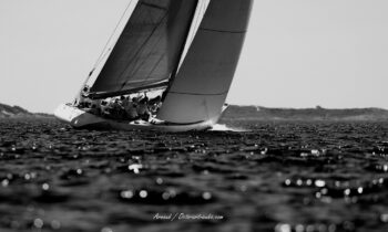 Force et lumière au ras des vagues. Le 12mJI French Kiss (F-12) sculptant la Méditerranée sous la gîte lors de l'Europa Cup 2026. Tirage d'art disponible en édition limitée. Photographie d'art en noir et blanc du voilier de tradition 12mJI French Kiss F-12 naviguant à forte gîte lors de l'Europa Cup 2026 à Hyères. Vue de trois-quarts arrière avec premier plan sur la mer scintillante.
