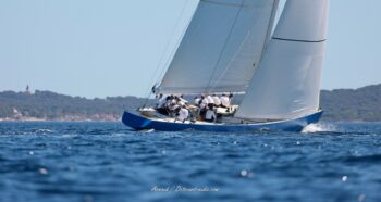 Cap sur les Îles d'Or. Le mythique 12mJI France 1 glisse majestueusement devant les côtes de Porquerolles lors de l'Europa Cup 2026. Tirage d'art disponible en édition limitée. Photographie de profil du voilier de tradition 12mJI France 1 à coque bleue naviguant toutes voiles dehors devant l'île de Porquerolles et son phare, lors de l'Europa Cup 2026 à Hyères.
