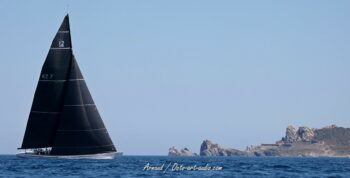 Rencontre de géants. Le New Zealand (KZ-7) glisse toutes voiles dehors devant le célèbre rocher du Crocodile à Porquerolles durant l'Europa Cup 2026. Tirage d'art disponible en édition limitée. Photographie de paysage marin montrant le voilier de course 12mJI New Zealand KZ-7 sous voiles noires naviguant devant le rocher du Crocodile à Porquerolles, lors de l'Europa Cup 2026.
