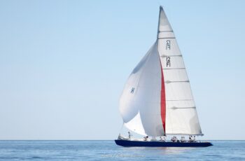 Majestueux portant en mer. Le mythique 12mJI France 1 déploie toute sa voilure et son spinnaker tricolore dans la rade de Hyères lors de l'Europa Cup 2026. Tirage d'art disponible en édition limitée. Photographie en plan large du voilier classique 12mJI France 1 à coque bleue naviguant de profil sous grand-voile et grand spinnaker blanc et rouge lors de l'Europa Cup 2026 à Hyères.