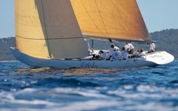 L'esprit de 1987 dans la baie. Le mythique 12mJI French Kiss (F-12) toutes voiles dehors lors de l'Europa Cup 2026 à Hyères. Tirage d'art disponible en édition limitée. Photographie de profil du voilier classique 12mJI French Kiss F-12 naviguant sous le soleil à l'Europa Cup 2026 à Hyères. Vue rasante sur la coque blanche et l'équipage dans le cockpit.