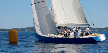 Éclat bleu roi sur la Méditerranée. Le mythique 12mJI France 1 et sa coque en bois emblématique à la relance après le passage de marque lors de l’Europa Cup 2026 à Hyères. Tirage d'art disponible en édition limitée. Proposition : Photographie de trois-quarts arrière du voilier de tradition 12mJI France 1 à coque bleue, glissant près d'une bouée jaune Toulon Provence Méditerranée lors de l'Europa Cup 2026 à Hyères. Vue sur l'équipage en action dans le cockpit.
