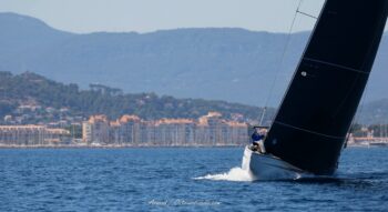 Retour au port sous haute tension. Un géant de la classe 12mJI glisse à vive allure devant les quais de Hyères à l'issue des épreuves de l'Europa Cup 2026. Photographie d'art disponible en tirage limité. Photographie de course d'un voilier classique de classe 12mJI naviguant à grande vitesse toutes voiles dehors devant les bâtiments du port de Hyères, lors de l'Europa Cup 2026. Vue sur la proue fendant le clapot et l'immense voilure noire sur fond de montagnes varoises.