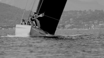Au cœur de la gîte. L'équipage du 12mJI New Zealand (KZ-7) fait corps avec la coque face aux vagues de la rade de Hyères lors de l'Europa Cup 2026. Tirage d'art graphique disponible en édition limitée. Photographie d'art en noir et blanc du voilier de tradition 12mJI New Zealand KZ-7 en pleine gîte pendant l'Europa Cup 2026 à Hyères. Vue arrière cadrée sur l'équipage au rappel et l'immense voile noire sur fond de reliefs côtiers.