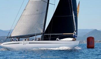 La légende Kiwi Magic dans les eaux varoises. Le mythique 12mJI New Zealand (KZ-7) à la marque de parcours lors de l'Europa Cup 2026 à Hyères. Photographie d'art disponible en tirage limité. Photographie de profil du voilier de tradition 12mJI New Zealand KZ 7 naviguant à proximité de la bouée orange de régate lors de l'Europa Cup 2026 à Hyères. Vue sur l'équipage aligné et la coque blanche aux lignes tendues face aux reliefs varois.