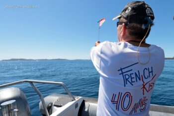 Dans les coulisses de la régate. Un officiel de l'organisation donne les signaux en mer, arborant le t-shirt anniversaire des 40 ans de French Kiss lors de l'Europa Cup 2026. Tirage d'art disponible en édition limitée. Photographie de dos d'un officiel en mer portant un t-shirt "French Kiss 40 ans" et levant un drapeau de signalisation depuis un bateau semi-rigide lors de l'Europa Cup 2026 à Hyères.