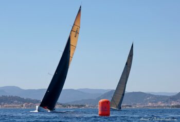 Duel tactique dans la rade. Le 12mJI France (F-7) mène la danse en pleine gîte après le passage de la bouée Métropole Toulon Provence Méditerranée lors de l’Europa Cup 2026. Tirage d'art disponible en édition limitée. Photographie de course au large mettant en scène le duel de deux voiliers classiques de la classe 12mJI, dont le France F-7 au premier plan en pleine gîte, contournant la bouée orange TPM lors de l'Europa Cup 2026 à Hyères.