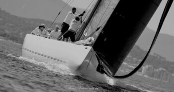 Rigueur et synchronisation. L'équipage du 12mJI France (F-7) à la manœuvre dans la brise hyéroise lors de l'Europa Cup 2026. Une esthétique intemporelle disponible en tirage d'art limité. Photographie artistique en noir et blanc du voilier classique 12mJI France F-7 naviguant à grande vitesse lors de l'Europa Cup 2026 à Hyères. Vue de trois-quarts avant sur l'équipage en action au pied du mât et l'écume sur la coque blanche.