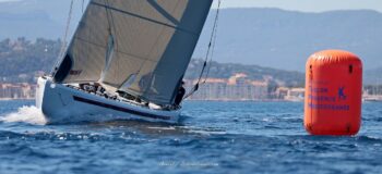 Cap sur la marque. Le 12mJI South Australia (KA-8) en approche frontale de la bouée Métropole Toulon Provence Méditerranée (TPM) sous le soleil de l’Europa Cup 2026. Tirage d'art disponible en édition limitée. Photographie de face du voilier de tradition 12mJI South Australia KA-8 approchant une bouée de régate orange gonflable Toulon Provence Méditerranée lors de l'Europa Cup 2026 à Hyères. La coque blanche fend l'eau avec les reliefs de la côte varoise en arrière-plan.