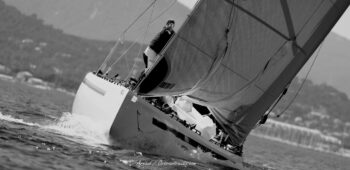 Lignes de fuite et puissance pure. La gîte spectaculaire d'un 12 Mètre International fendant le plan d'eau hyérois sous la brise de l'Europa Cup 2026. Tirage d'art graphique disponible en édition limitée. Photographie artistique en noir et blanc d'un voilier classique de classe 12mJI en pleine gîte lors de l'Europa Cup 2026 à Hyères. Vue arrière sur l'écume, la coque inclinée et un équipier au rappel face aux côtes du Var.