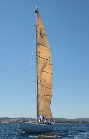 Intervention de haute voltige au sommet du mât du 12mJI France (F-7) lors du retour au port. Un instantané de la rigueur et de la technique requises à bord de ces géants de la Coupe de l'America pendant l'Europa Cup 2026. Photographie de profil du voilier classique 12mJI France F-7 en mer près de Hyères, avec un équipier hissé en haut du mât pour une intervention technique. Vue sur la grand-voile couleur ambre et la coque blanche sur fond de côtes varoises.
